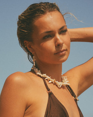 Model wearing a brown bikini with a pearl necklace against a clear blue sky.