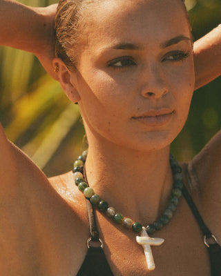 Model with a necklace featuring a cross and beads against a blurred natural background