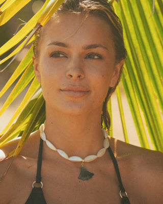 Model wearing a brown bikini with a shell necklace, standing under palm leaves.