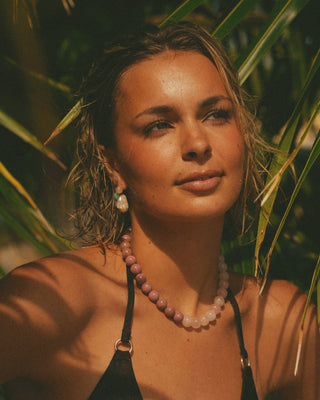 Model with a pink necklace and earrings standing among palm leaves