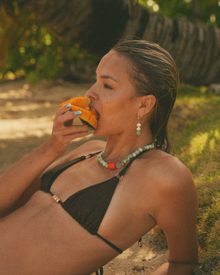 Model in a brown bikini wearing coral necklace sitting outdoors, holding a papaya.